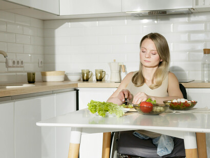 A woman in a beige tank top sitting in her kitchen near a table laid with vegetables