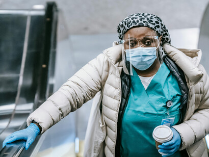 A masked Black nurse rides the escalator in the underground, quietly overlooked by everyone passing by