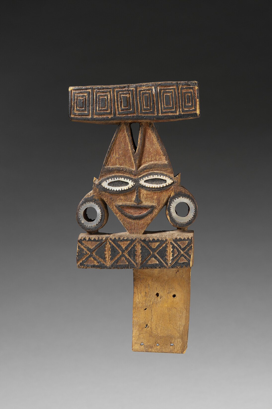 Sculpture monoxyle représentant un visage stylisé encadré par deux rectangles de bois gravés de motis géométriques. Le visage central est en forme de losange dont la partie supérieure se sépare elle-même en deux triangles. Les yeux comme les lobes des oreilles sont incrustés de nacre. De la peinture noire rehausse les traits du visage et les motifs géométriques au haut et bas de la figure. Dans la partie inférieure, un tenon plat en bois (fragmentaire) prolonge la sculpture permettant la fixation de l'objet au canot © musée du quai Branly, photo Patrick Gries, Valérie Torre