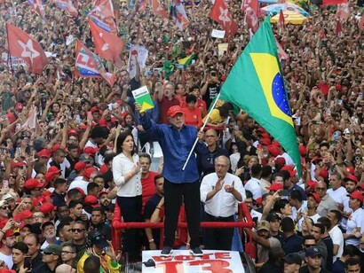 Visto em um plano mais aberto, Lula aparece sobre uma plataforma elevada, acompanhado por aliados e cercado por uma multidão que se estende por toda a imagem. Bandeiras brasileiras e do Partido dos Trabalhadores se misturam entre os presentes, revelando a dimensão coletiva da mobilização. Mais do que a presença de um líder, a fotografia mostra a força de uma base social que se reúne em torno de um projeto político. No artigo, Boaventura de Sousa Santos trata a vitória eleitoral como parte de uma disputa mais ampla sobre os rumos do Brasil e do mundo. A questão decisiva, nessa perspectiva, é como converter a energia demonstrada nas ruas em participação democrática, inclusão social e mudanças duradouras