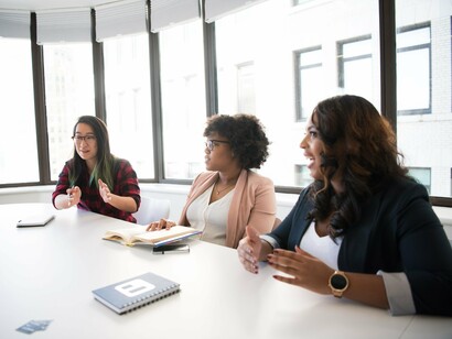 Três mulheres participam de uma conversa em um escritório, voltadas umas para as outras. Mais do que transmitir informações, a linguagem permite aos interlocutores interagir e produzir sentidos em uma determinada situação social