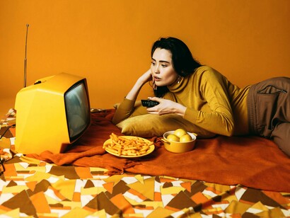 Mulher se distrai em frente à televisão. Eu cresci, ainda longe da vida dos sonhos, mais abracei cada nova fase, com amor, com ódio, medo, alegria e tristeza, faz parte assim como a arte, sempre foi uma forma de terapia para mim, meu combustível secreto para não desistir e lembrar de quem sou