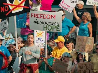 A diverse crowd of protesters raising handmade signs in a public demonstration, representing popular resistance to the concentration of political power
