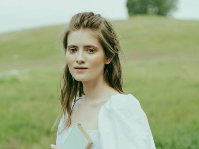 A young woman holding a book close to her chest, standing against a backdrop of rolling green hills, reminds us how the digital age has expanded our horizons but often leaves us searching for deeper understanding and meaning in a rapidly changing world