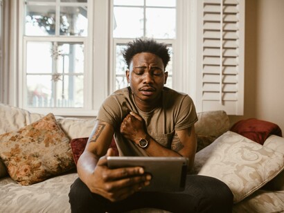 A man sitting on a couch, looking at his tablet with an emotional expression