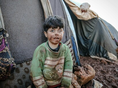 A young child sits in the mud outside a worn-out tent, his face and clothes covered in dirt—symbolizing the harsh reality of those left behind by failed aid efforts