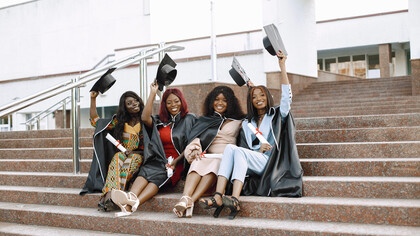 A picturesque scene unfolds as a group of young African American female students dons black graduation gowns against the backdrop of a campus setting