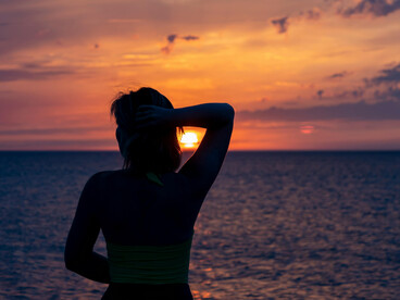 Woman standing by the sea at sunset, seen from behind