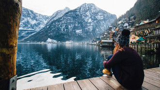 A woman sits on a wooden pier, gazing out at a serene winter landscape of a lake and snow-capped mountains