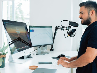 In a dynamic social media agency, a man in a black t-shirt works at a table, his workstation includes a desk with two screens, a prominent podcast microphone, and a computer with a splitscreen interface