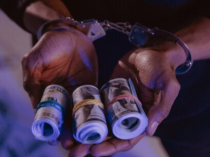 A pair of handcuffed hands clutches rolled bundles of hundred-dollar bills, evoking the inescapable grip of corruption that now defines the American power structure 