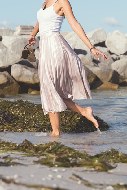 A woman stands gracefully on the beach, adorned in a white skirt and holding black leather flat sandals, exuding a sense of serenity and connection with the natural beauty of the seaside