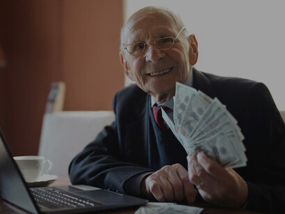 A joyful senior businessman, seated at his desk, effectively managing his laptop tasks while confidently handling a stack of money