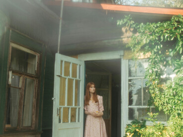 A young woman in a vintage dress pauses at the entrance of a rustic country house, enveloped by rich greenery, blooming flowers, and gentle summer light