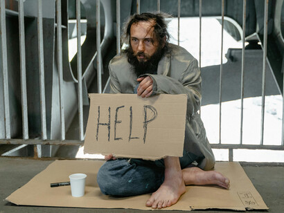 A male beggar is sitting on the ground, holding a sign that says “Help”
