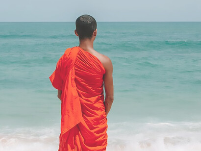 A man, adorned in Buddhist garments, stands contemplatively on the seashore