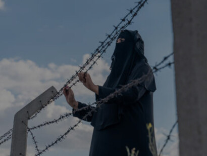 Behind a chain-link fence, a refugee woman gazes out, her expression a mix of hope and apprehension