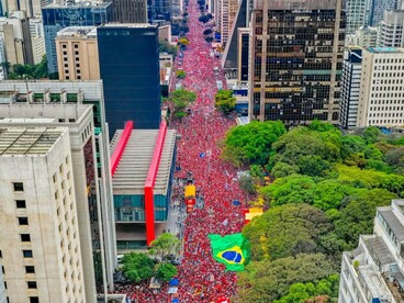 Vista de um ponto elevado, uma multidão ocupa uma extensa avenida cercada por edifícios altos e áreas arborizadas. Uma grande bandeira brasileira destaca-se no meio da concentração, que dá expressão visual à mobilização popular e à disputa pelo futuro político do país