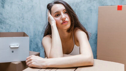 A woman, encircled by boxes, wears a somber expression, reflecting her emotions as she prepares for a move