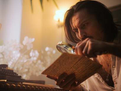 Focused man studying an old document through a magnifying glass in a warmly lit vintage workspace, evoking themes of investigation, discovery, and historical inquiry