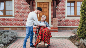 A man assisting an elderly person in a wheelchair in front of a house