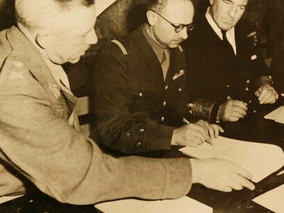 La resa della Germania, 7 maggio 1945. Il Generale Francois Sevez, rappresentante del Generale Alphonse Juin, Capo di Stato Maggiore del Generale de Gaulle, appone la sua firma sul documento con il quale tutta la resistenza tedesca rimanente capitola. Alla sua destra il Tenente Generale Sir F.E. Morgan, Vice Capo di Stato Maggiore, e alla sua sinistra l'Ammiraglio Harold M. Burrough, Comandante in Capo delle Forze Navali di Spedizione Alleate