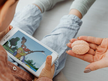 A child is engrossed in phone play while seated beside a parent who holds a brain-shaped toy