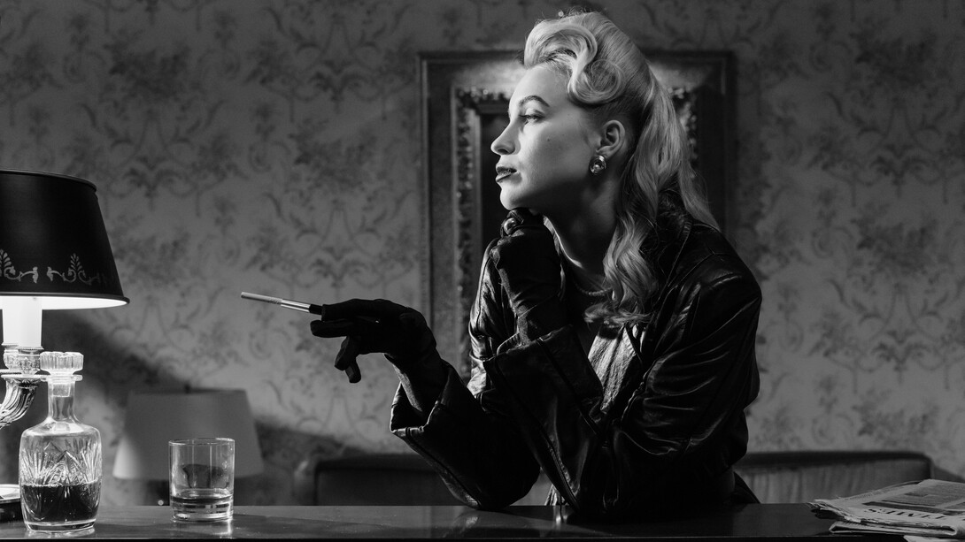 Atriz vestida de um figurino de couro em cena enigmática tramando os próximos passos de seu plano