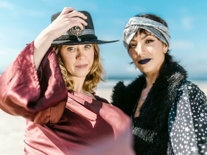 Duas mulheres sentadas na areia, com looks boho e acessórios marcantes, exploram a estética livre e criativa típica dos festivais onde estilo e personalidade caminham juntos na experiência