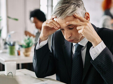 An exhausted businessman, head down on his desk, illustrates the toll of a toxic work environment as stressed colleagues in the background navigate anxiety, conflict, and dissatisfaction