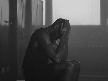 A male athlete sits alone in the locker room, visibly stressed and overwhelmed