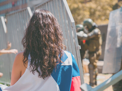 Joven mujer con una bandera de chile a sus espaldas encara una barricada de los carabineros de la policía chilena en Santiago de Chile