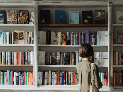 A woman wearing a beige long-sleeve shirt stands before a white wooden bookshelf