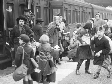 British children waiting on a train platform with small suitcases and gas masks slung over their shoulders during World War II, UK