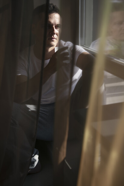 A contemplative man sits alone near a window, with soft natural light and curtain shadows creating an introspective atmosphere