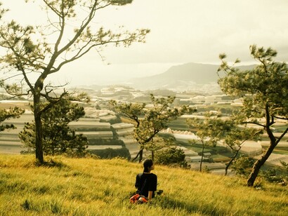 A person resting on a hill between nature and the city, gazing outward in thoughtful silence