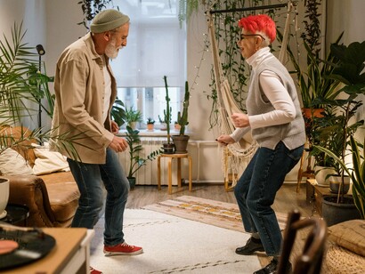 Um casal idoso dança em meio às plantas de uma sala, em um momento descontraído dentro de casa. A cena sugere que novas experiências e formas de viver não precisam obedecer a uma idade ou a um roteiro previamente estabelecido