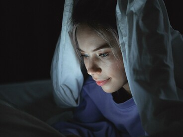 Sob o cobertor, a mulher utiliza um tablet durante a noite. O quarto, tradicionalmente associado ao repouso, também se transforma em espaço de consumo de conteúdo e prolongamento do tempo diante das telas