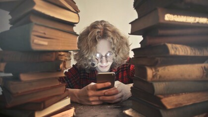 A young boy scrolling, glued to his smartphone while ignoring the piles of old books in front of him; an image that reflects the reality of the youth in our current generation, all about their smartphones and movie platforms, rejecting the good old novels