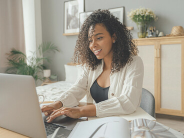 A woman in a white blazer sits at a brown wooden table, working on a MacBook Pro