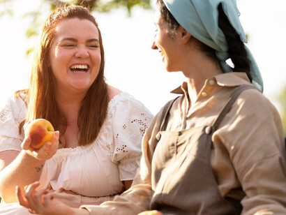 Two smiling women sit together on a sunny patio, surrounded by a colorful spread of fresh fruits, enjoying a delightful moment of friendship