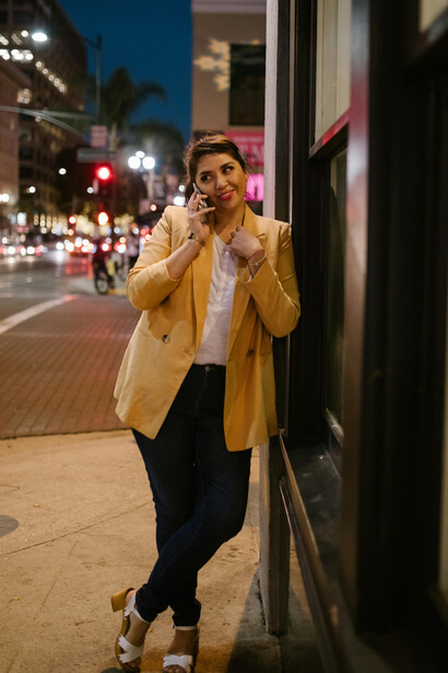 At night, a woman in a beige coat and black pants stands by a building, speaking on the phone, suggesting a long-distance connection