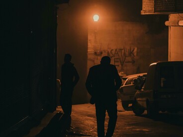 Silhouettes of men move along a narrow road as figures drift across a dark, fog-covered pavement