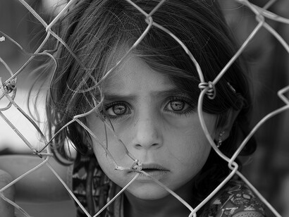 A young girl's piercing eyes peer through a fence, reflecting the innocent cost of civilization's moral and political collapse