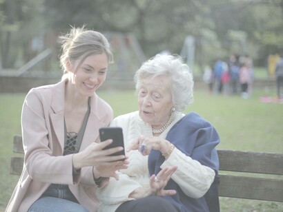 An elderly woman marveling at the latest technology in a park with her younger relative