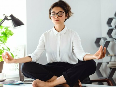 A woman meditates on her desk, surrounded by paperwork, representing the struggle for balance in the energy–work equation, where professionals seek relief from workplace pressures