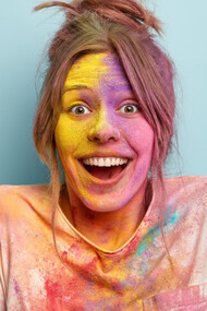 Headshot of an optimistic woman in a white t-shirt, smiling as she plays with vibrant colors, her face and clothes covered in colorful splashes