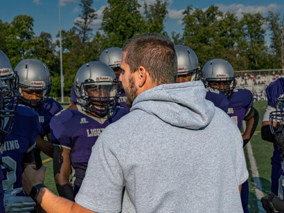 A coach wearing athletic gear shouts instructions to players during an American football practice