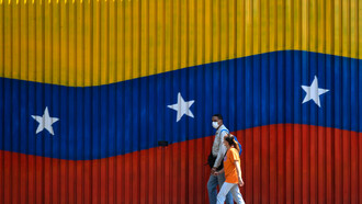 Una pareja camina frente a una barda decorada con la bandera de Venezuela