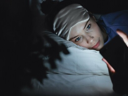 Na cama, a mulher manuseia o celular enquanto permanece envolvida pela atmosfera silenciosa da noite. Desligar-se das redes também significa criar espaço para que o corpo e a mente desacelerem